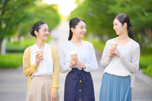 3人の女性が紙コップを持って歩きながら笑顔で会話している公園の風景